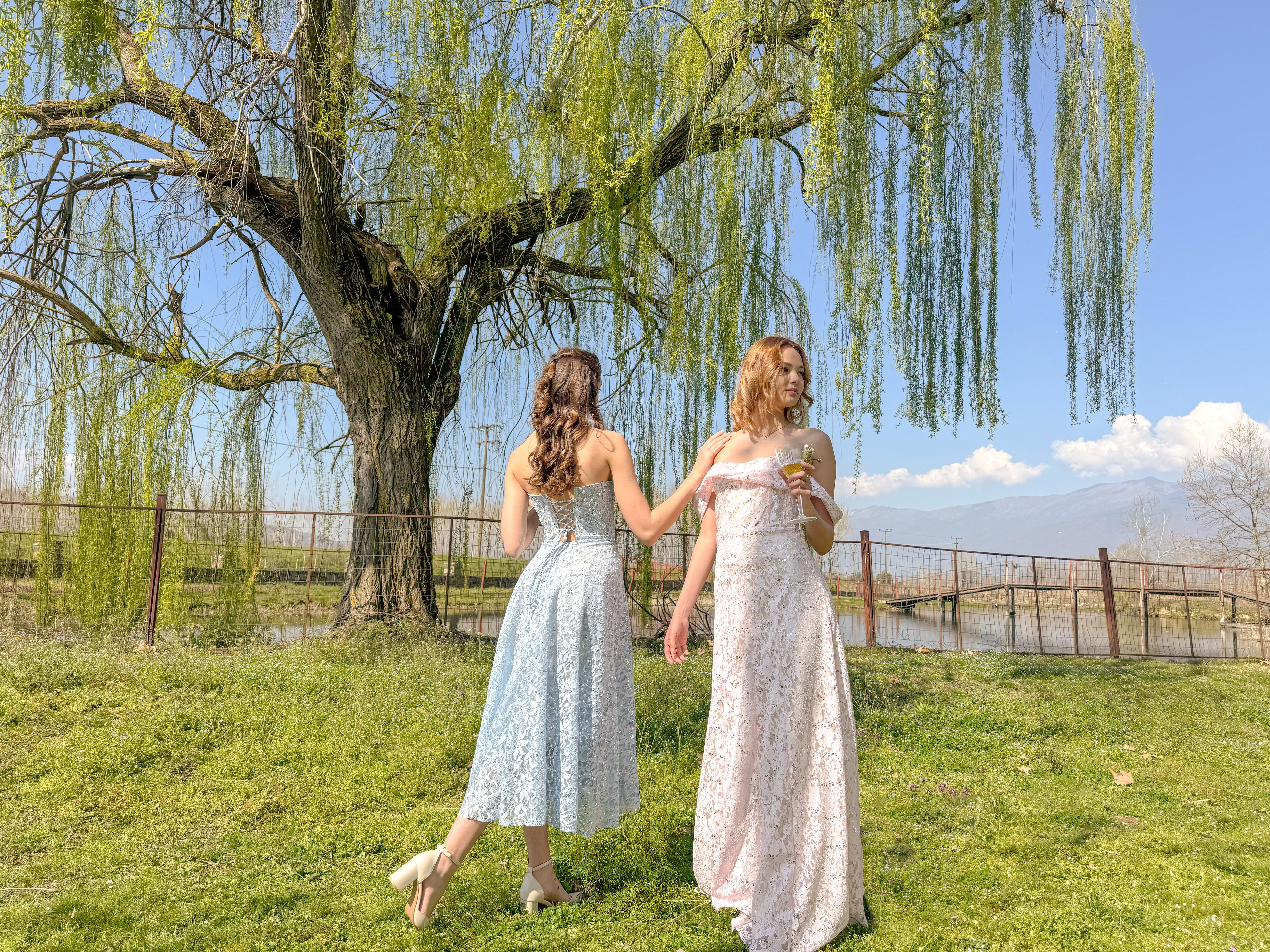 Two women in white dresses standing under a willow tree by a lake.