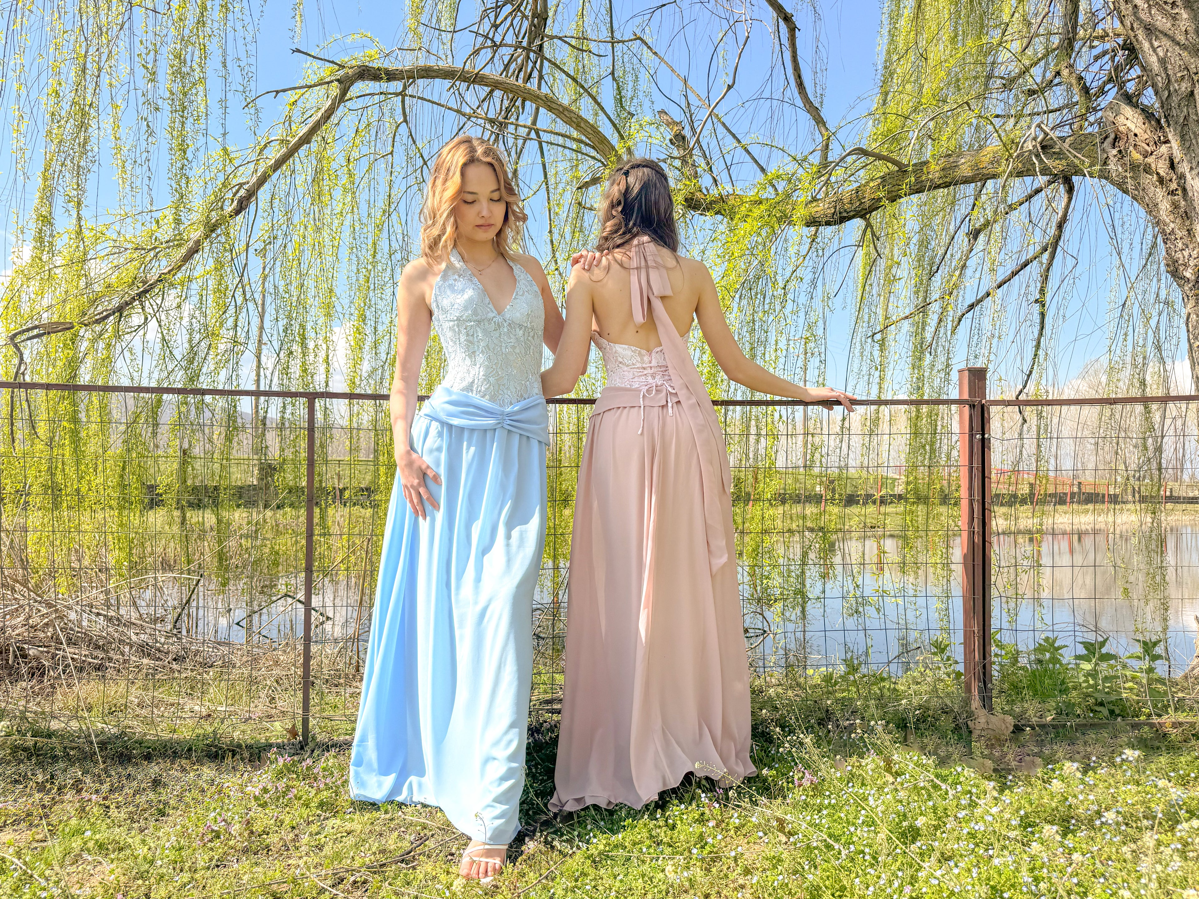 Two women in long dresses standing by a fence with a scenic background
