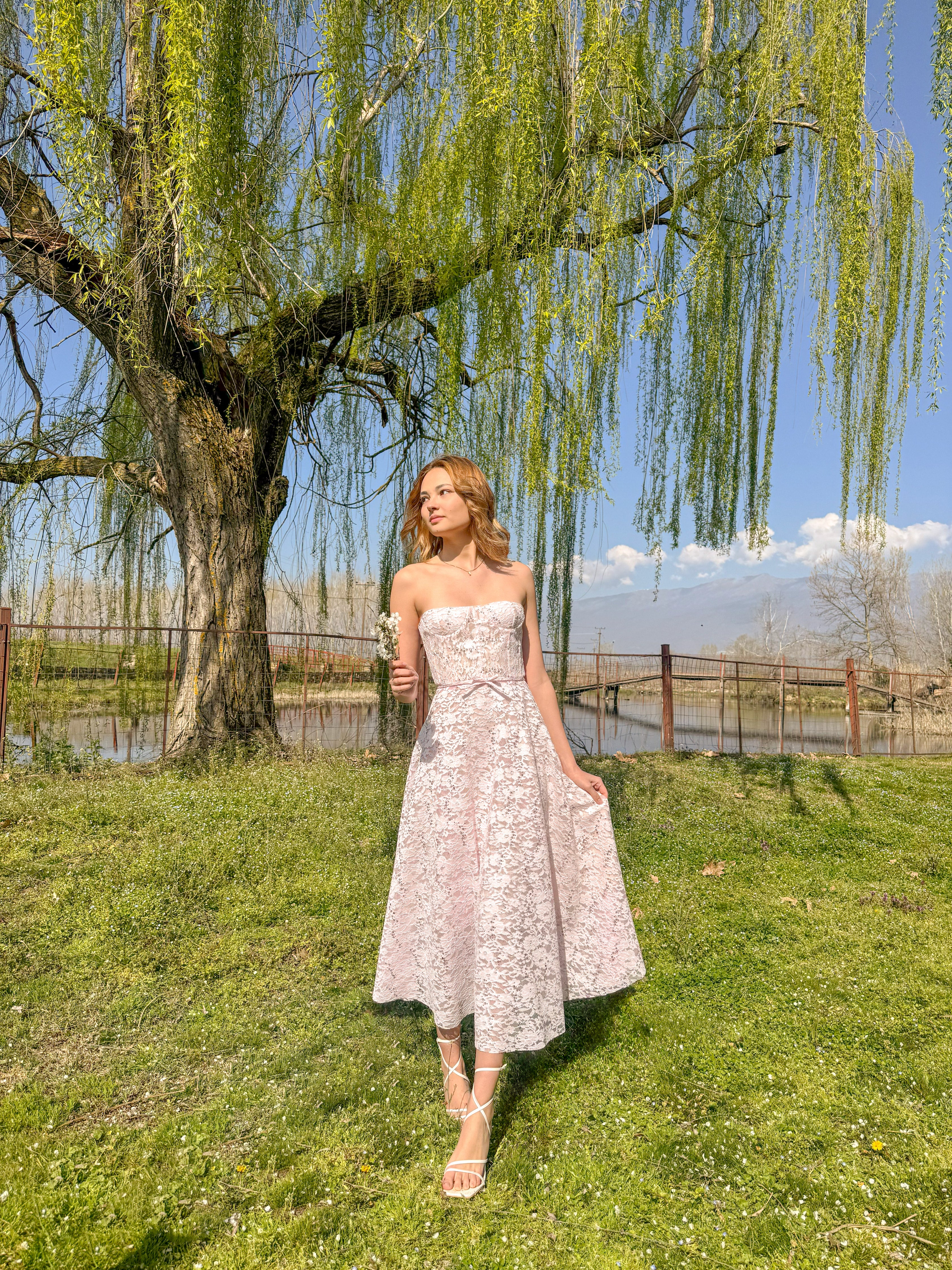 Woman in a strapless floral dress standing under a willow tree by a lake.